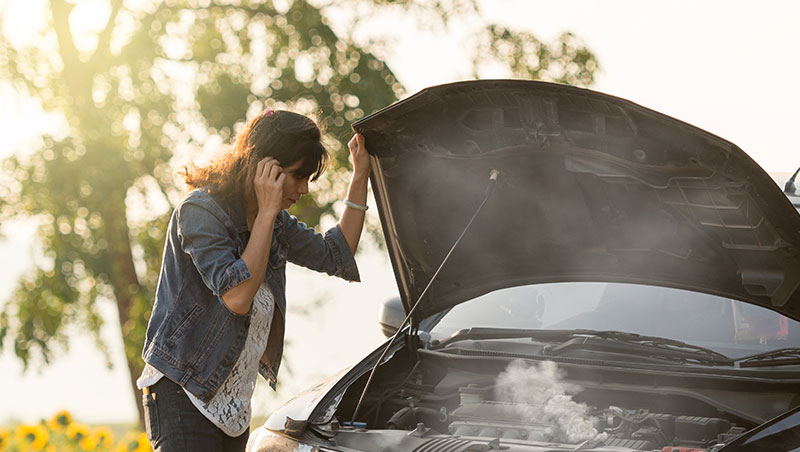 A photo depicting a person next to their car, the hood lifted, revealing smoke coming from the engine